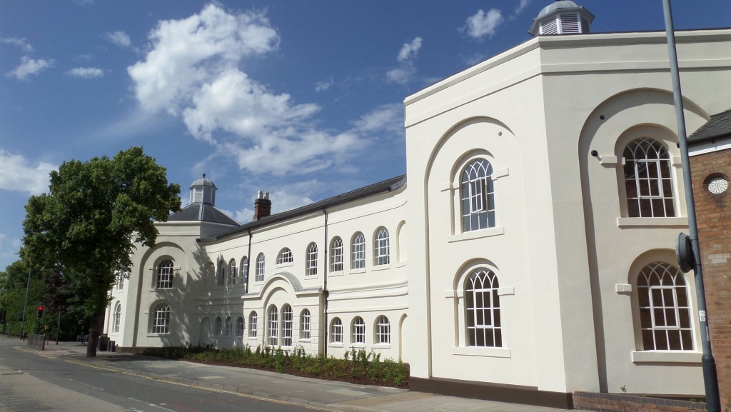 Cream-colored building with arched windows and decorative circular arches under a bright blue sky. A tree lines the street to the left.